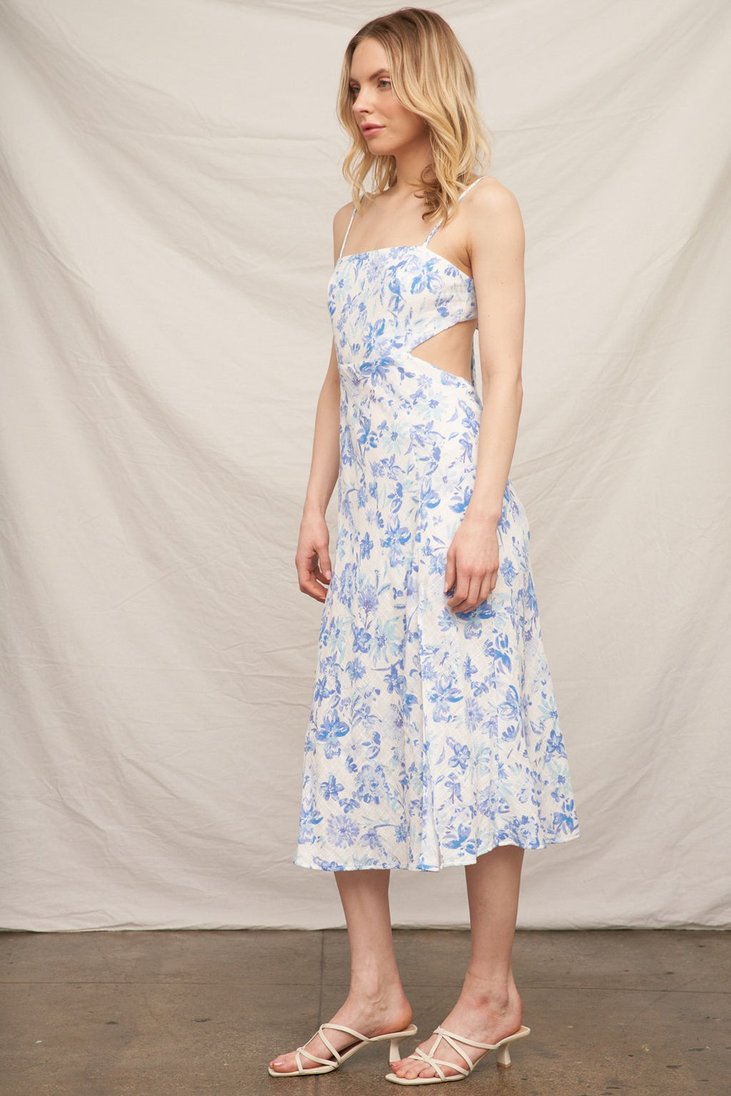 Blue floral spaghetti-strap square-neck side-cut-out back-bow-tie front-slit midi dress - side three-quarter view showing spaghetti straps, open side cut-out at waist profile, fluid A-line midi skirt silhouette, and blue floral print on white from the side