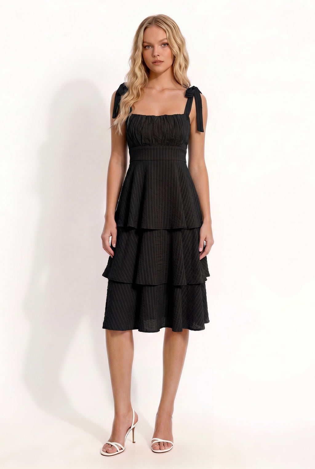 Black shoulder-tie-strap ruched-bust smocked-waist three-tier midi dress - front three-quarter angled view showing shoulder tie bows, ruched bust, smocked waist, and three-tier skirt silhouette from the side in deep black crinkle