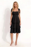 Black shoulder-tie-strap ruched-bust smocked-waist three-tier midi dress - front three-quarter angled view showing shoulder tie bows, ruched bust, smocked waist, and three-tier skirt silhouette from the side in deep black crinkle