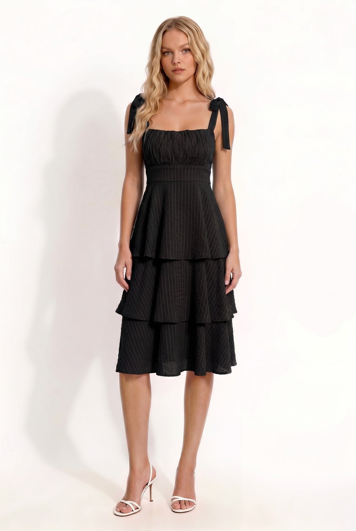 Black shoulder-tie-strap ruched-bust smocked-waist three-tier midi dress - front three-quarter angled view showing shoulder tie bows, ruched bust, smocked waist, and three-tier skirt silhouette from the side in deep black crinkle