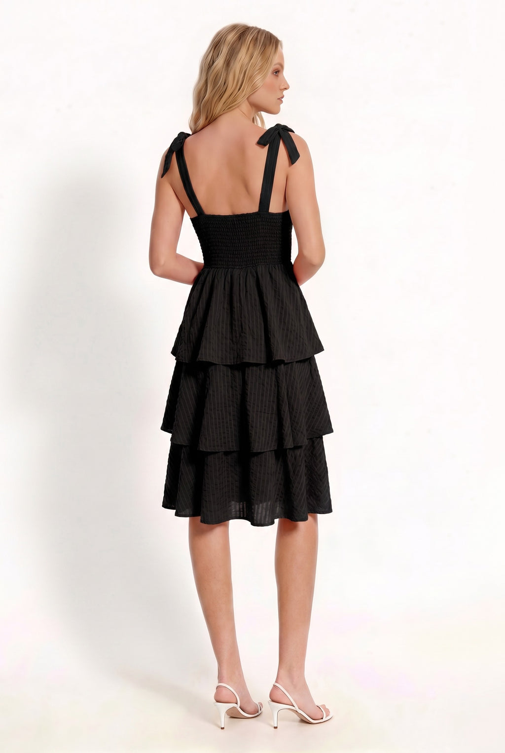 Black shoulder-tie-strap ruched-bust smocked-waist three-tier midi dress - side three-quarter view showing wide strap with shoulder bow, ruched bust profile, smocked waistband, and cascading three-tier midi skirt from the side in deep black