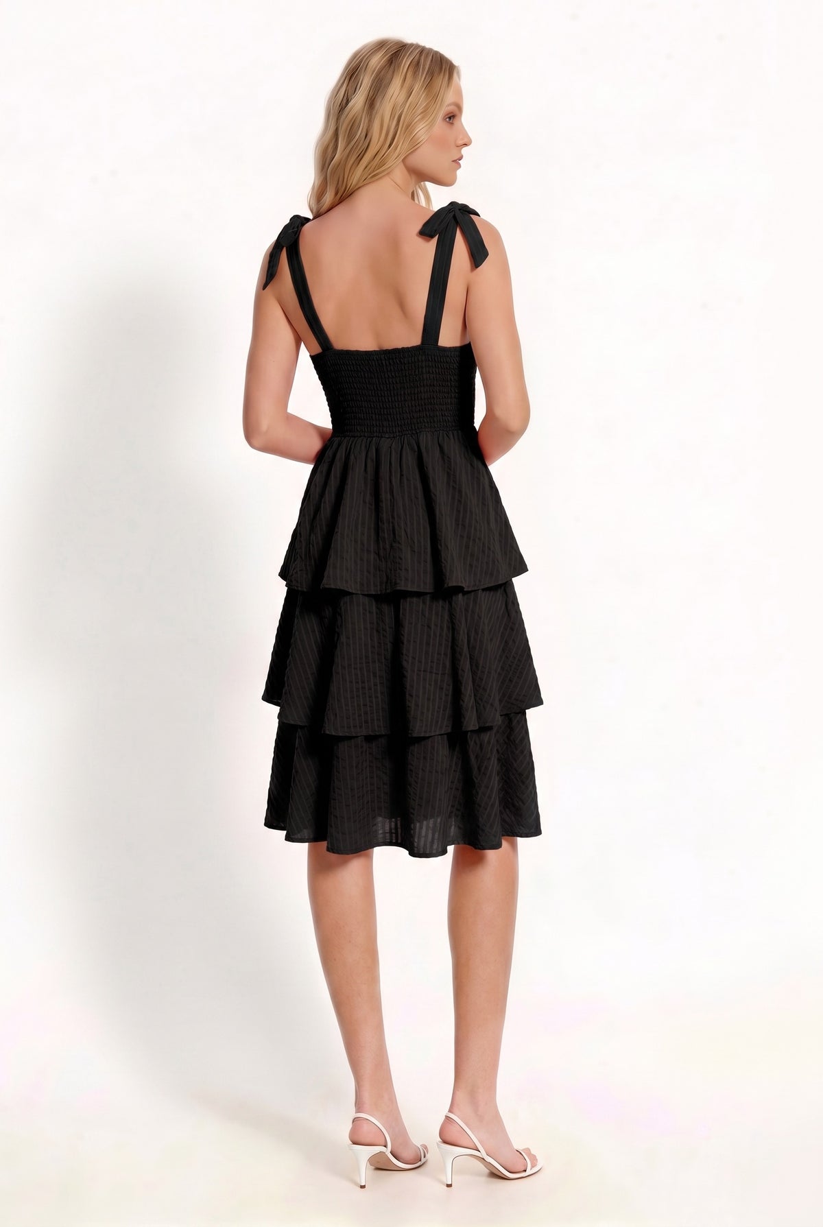 Black shoulder-tie-strap ruched-bust smocked-waist three-tier midi dress - side three-quarter view showing wide strap with shoulder bow, ruched bust profile, smocked waistband, and cascading three-tier midi skirt from the side in deep black