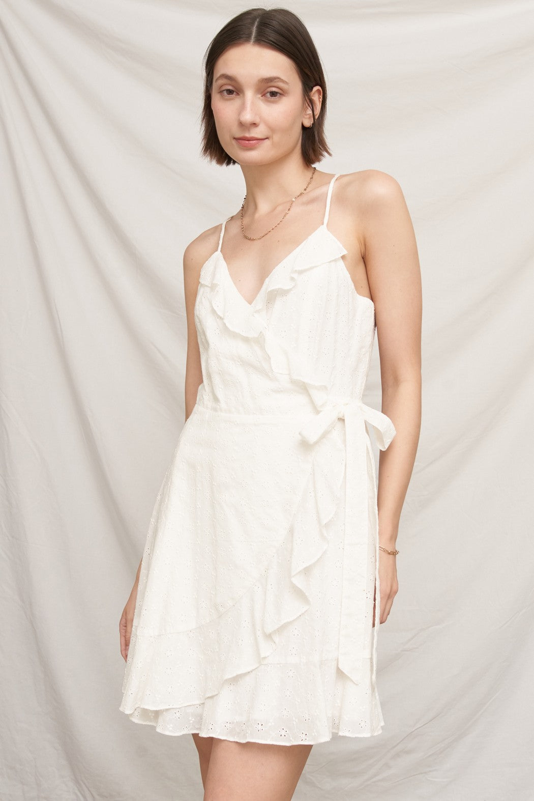 White eyelet spaghetti-strap V-neck ruffle-wrap tie-waist mini dress - third front three-quarter view showing ruffle wrap diagonal, waist tie bow, flared skirt silhouette, and full eyelet broderie anglaise hem
