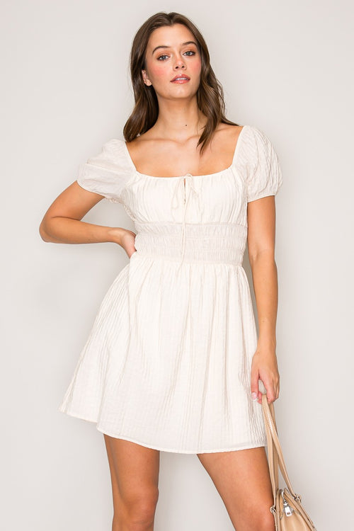 Cream textured square-neck front-tie-detail puff-sleeve smocked-waist babydoll mini dress - three-quarter side view showing puff sleeves, smocked waistband, and full skirt