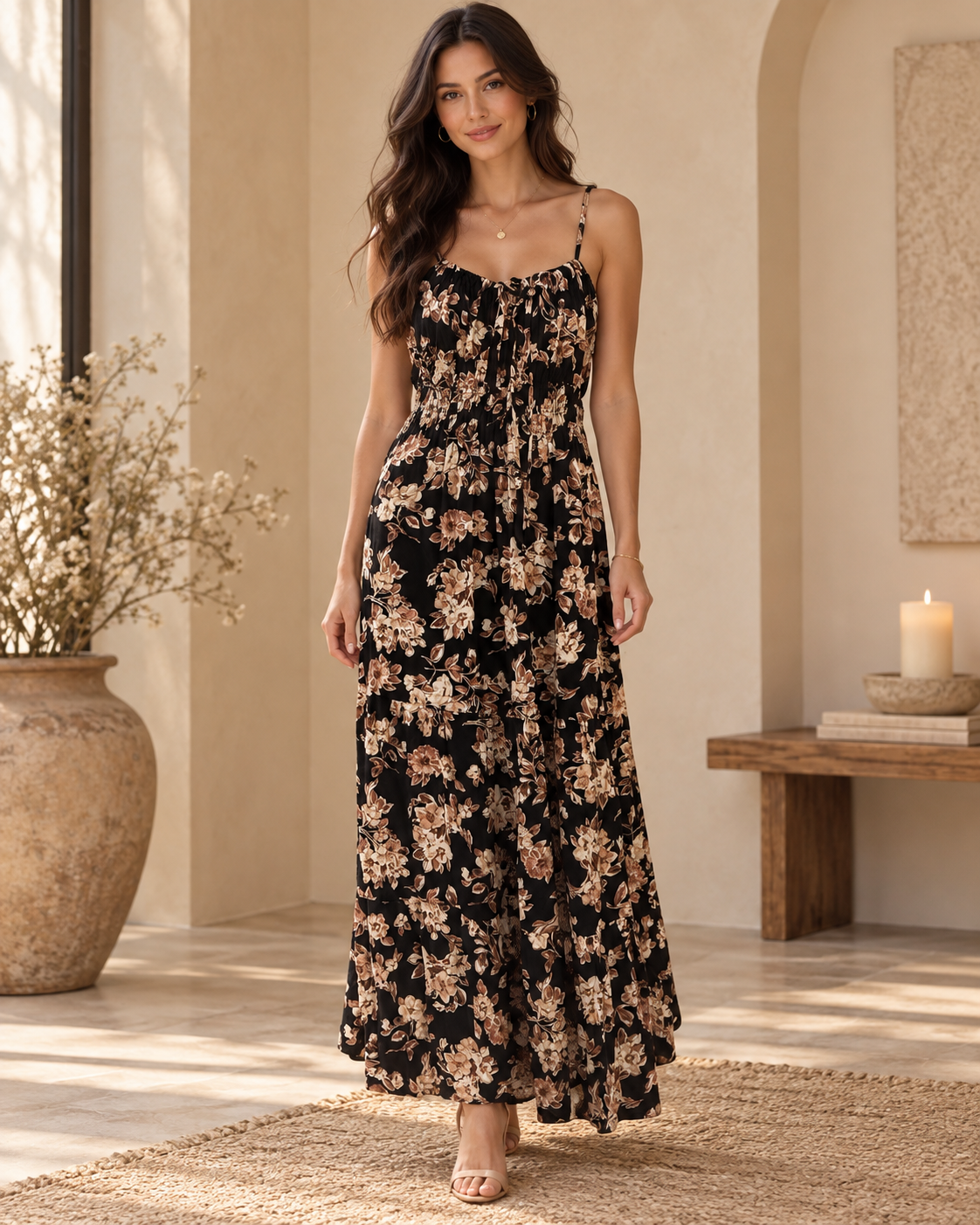 Black botanical-floral spaghetti-strap tie-front smocked-waist tiered maxi dress - front full-length lifestyle view showing fine spaghetti straps, centre-front tie bow neckline, smocked waistband, and voluminous two-tier floral chiffon maxi skirt in black botanical print with gold heels in warm interior setting